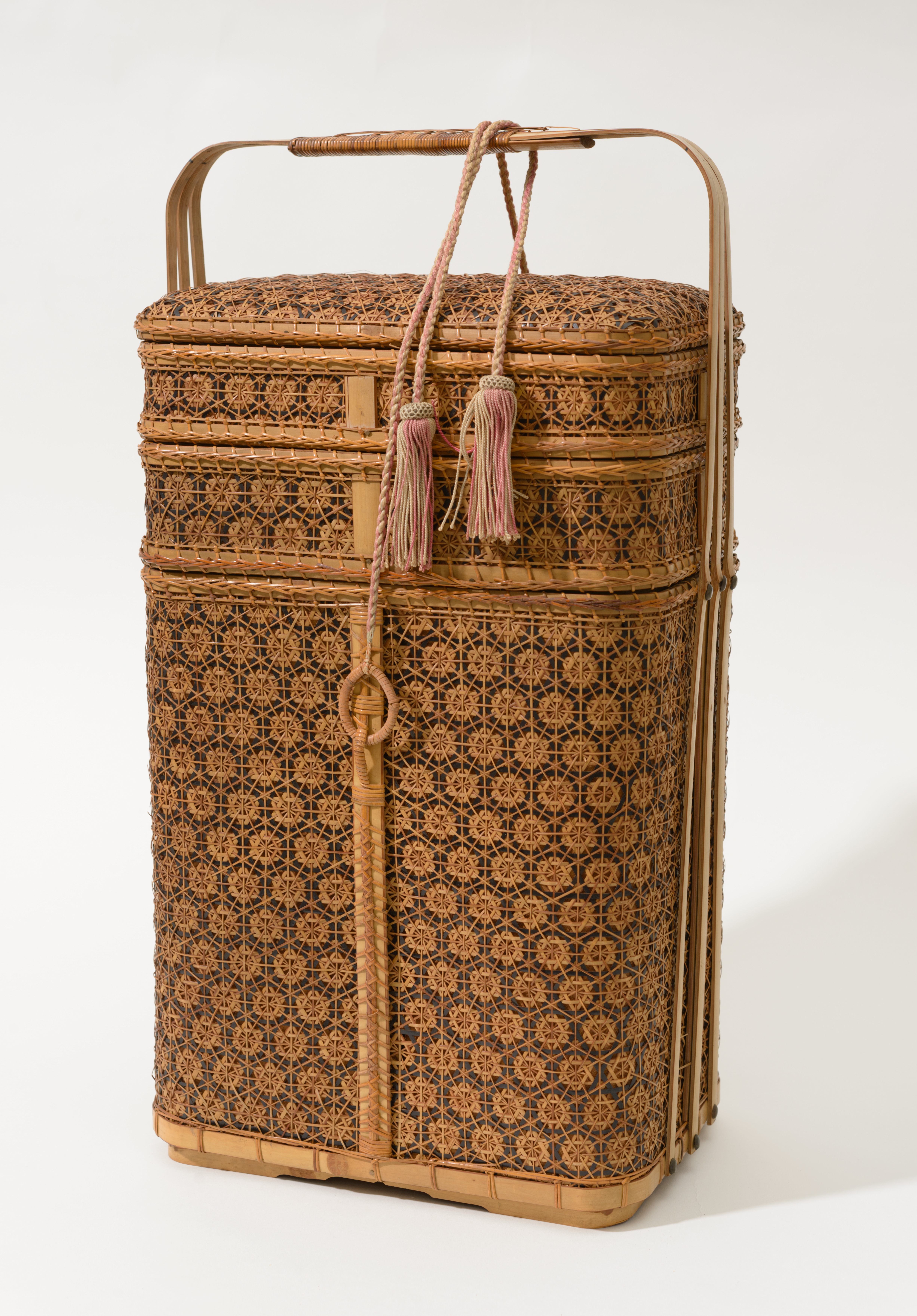 Three-tiered woven bamboo basket, with the silk tie reinstated to the repaired attachment loop. The tassel ties have been displayed looped and draped around the single handle which extends from the sides over the top of the basket.